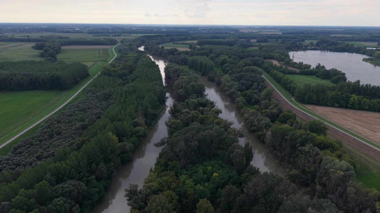 Aerial view of a narrow winding river cutting through dense forest and open farmland. The lush green trees contrast with the cultivated fields, creating a peaceful rural landscape under a cloudy sky