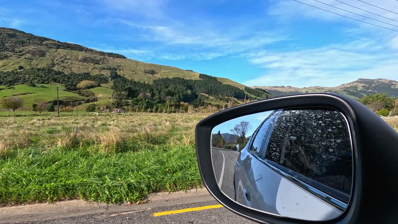 A moving car’s side mirror reflects scenic rural hills, roadside greenery, and blue skies in Akaroa, New Zealand, captured in bright daylight with smooth camera motion
