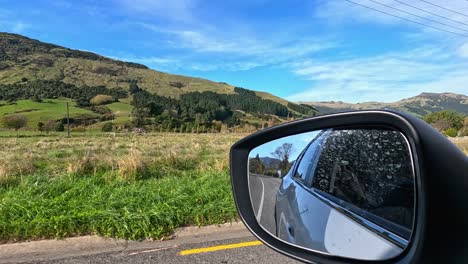 a moving car’s side mirror reflects scenic rural hills, roadside greenery, and blue skies in akaroa, new zealand, captured in bright daylight with smooth camera motion