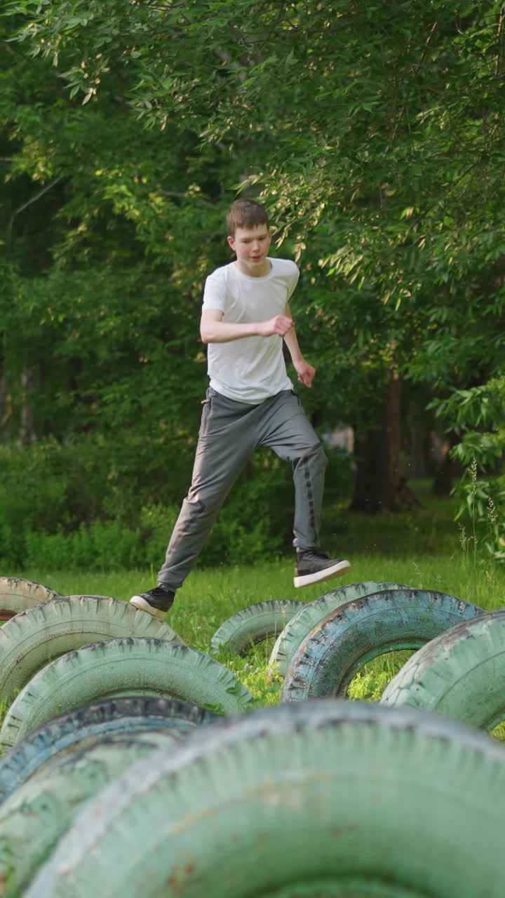 vista frontal de un niño en una camisa blanca y pantalones grises entrenamiento en una serie de neumáticos en un campo de hierba, con árboles y vista borrosa de la construcción del fondo