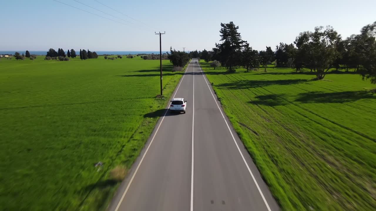 Drone view of white car driving along long straight road flanked by lush green fields and scattered trees under clear daylight sky in a peaceful rural setting