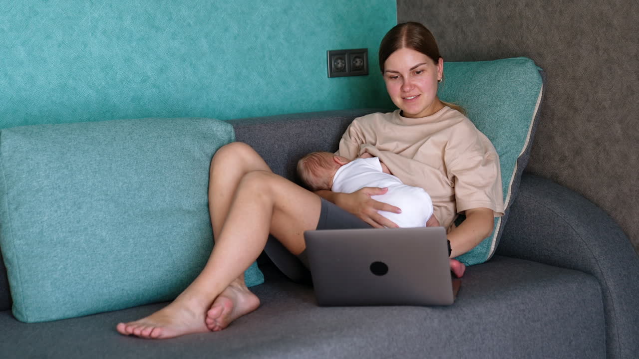 Young mom breastfeeds her newborn sitting on the sofa. Woman looks at laptop and works on it.