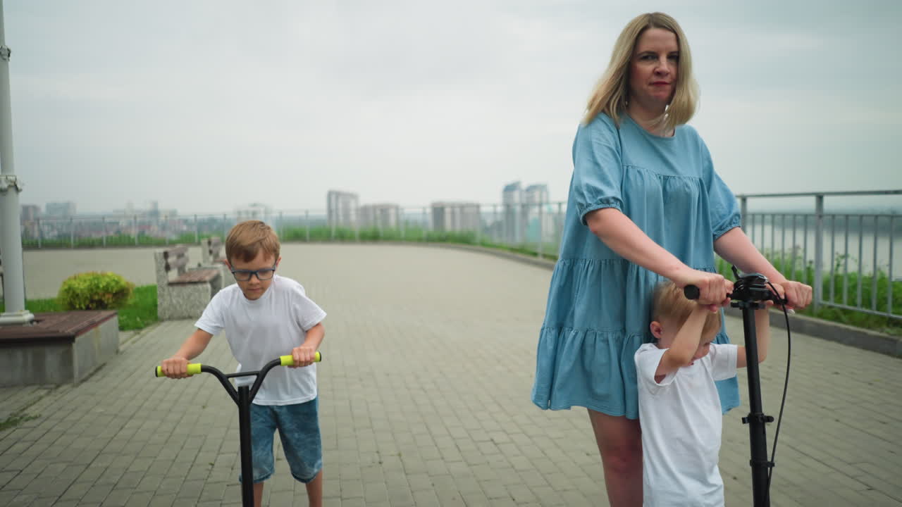женщина и ее двое детей ездят на скутерах на открытом воздухе по асфальтированной тропе с отдаленным городским пейзажем за ними