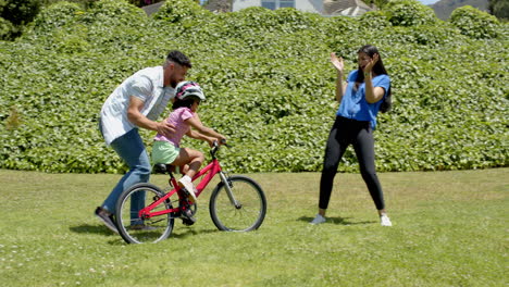 father teaching daughter to ride bicycle while mother clapping in park