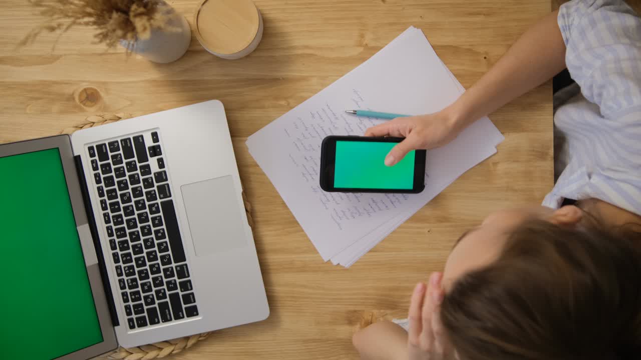 The girl looks at the phone with a green screen. Opposite is an open laptop with a green screen. The concept of remote work from top view