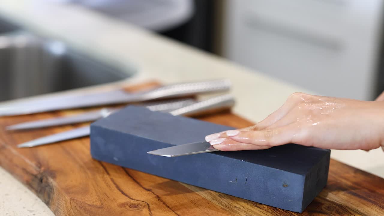 A person sharpens a knife on a whetstone in a well-lit kitchen, focusing on technique and precision