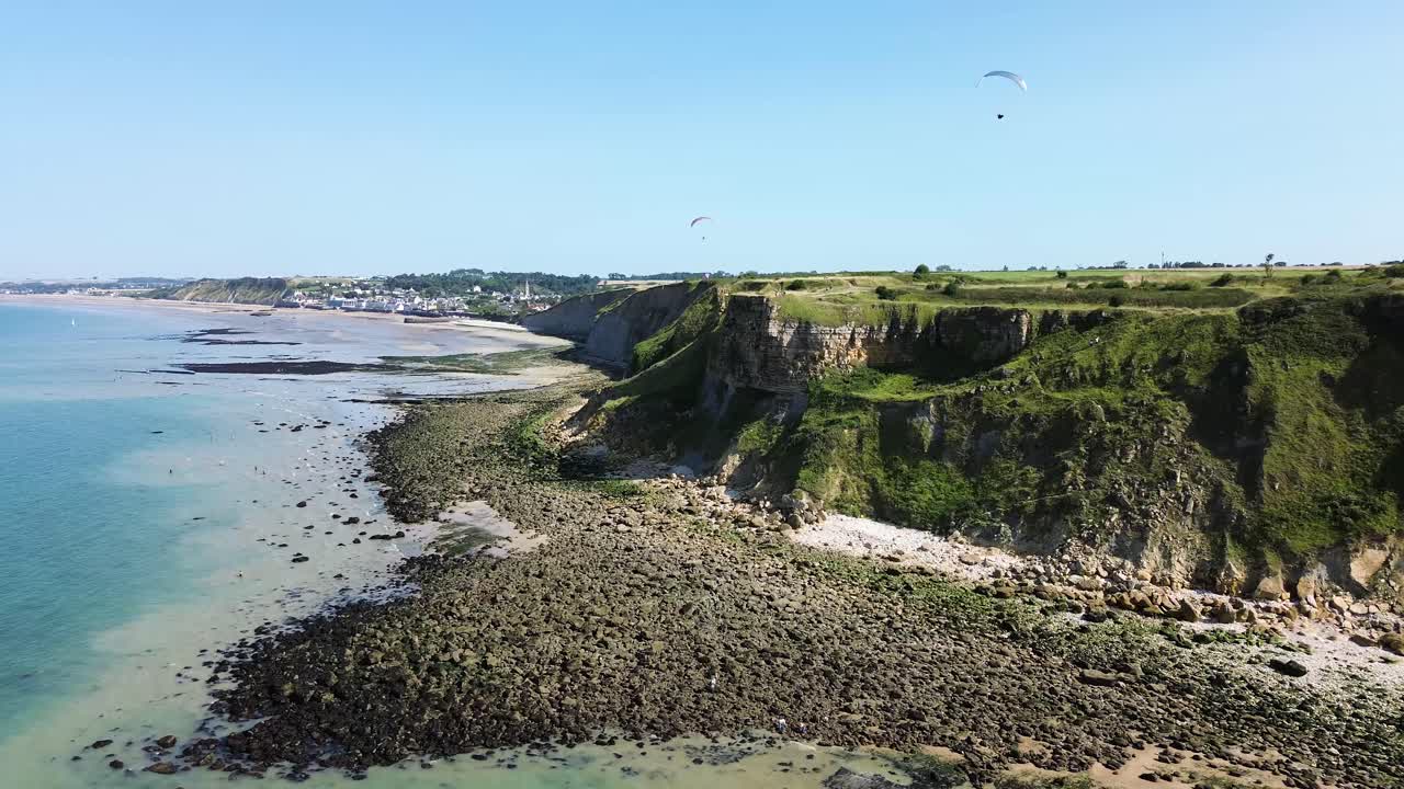 Drone footage gliding along the Arromanches cliffs, showing their majestic drop into the blue sea, framed by lush green fields under a cloudless sky