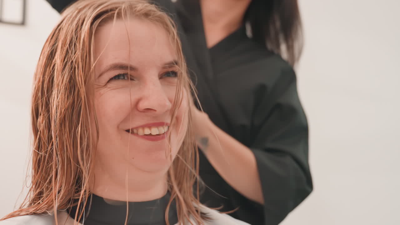 Donna che si fa applicare extension di trecce con perline in un salone, stilista che lavora sui capelli bagnati, cliente con mantellina, primo piano di un viso sorridente, trecce texturizzate, mani precise, illuminazione calda da studio, atmosfera calma e sicura