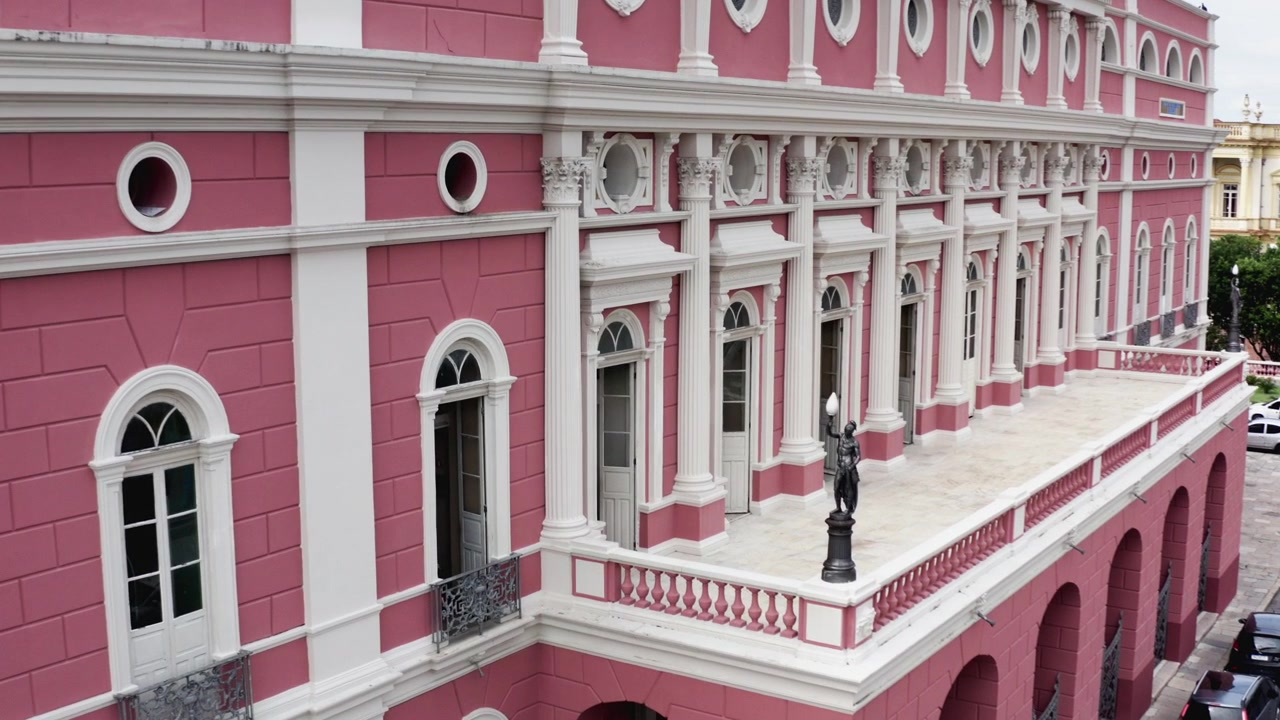 Drone avança ao longo da fachada lateral do Teatro Amazonas com paredes cor-de-rosa, arcadas brancas e janelas em arco até a balaustrada de canto e seu lampião com estátua de bronze, dia nublado, Manaus, Brasil 4K