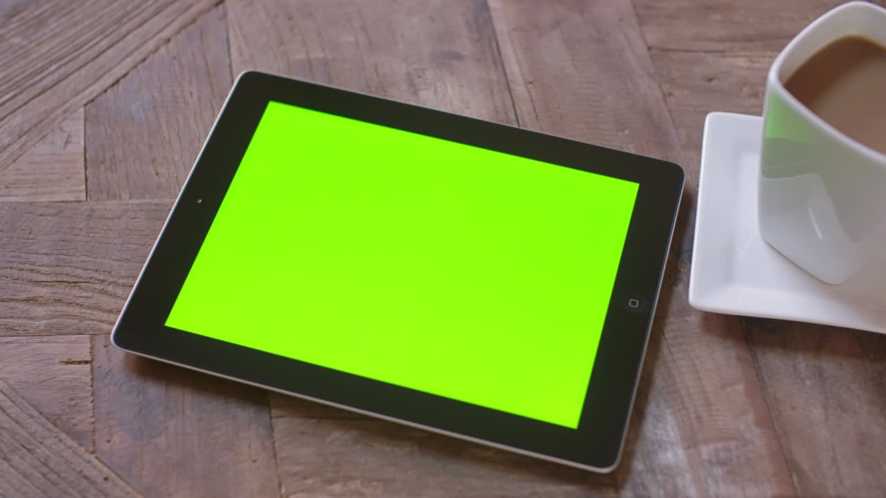 Ser ned på en tablet, der bevæger sig til højre, og en kaffekop på et bord med grøn baggrund.