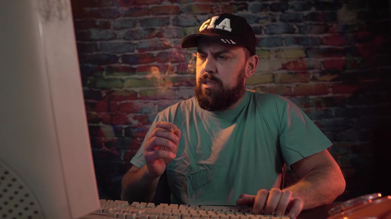 Datový analytik CIA kouří cigaretu u stolu před starým počítačovým monitorem.