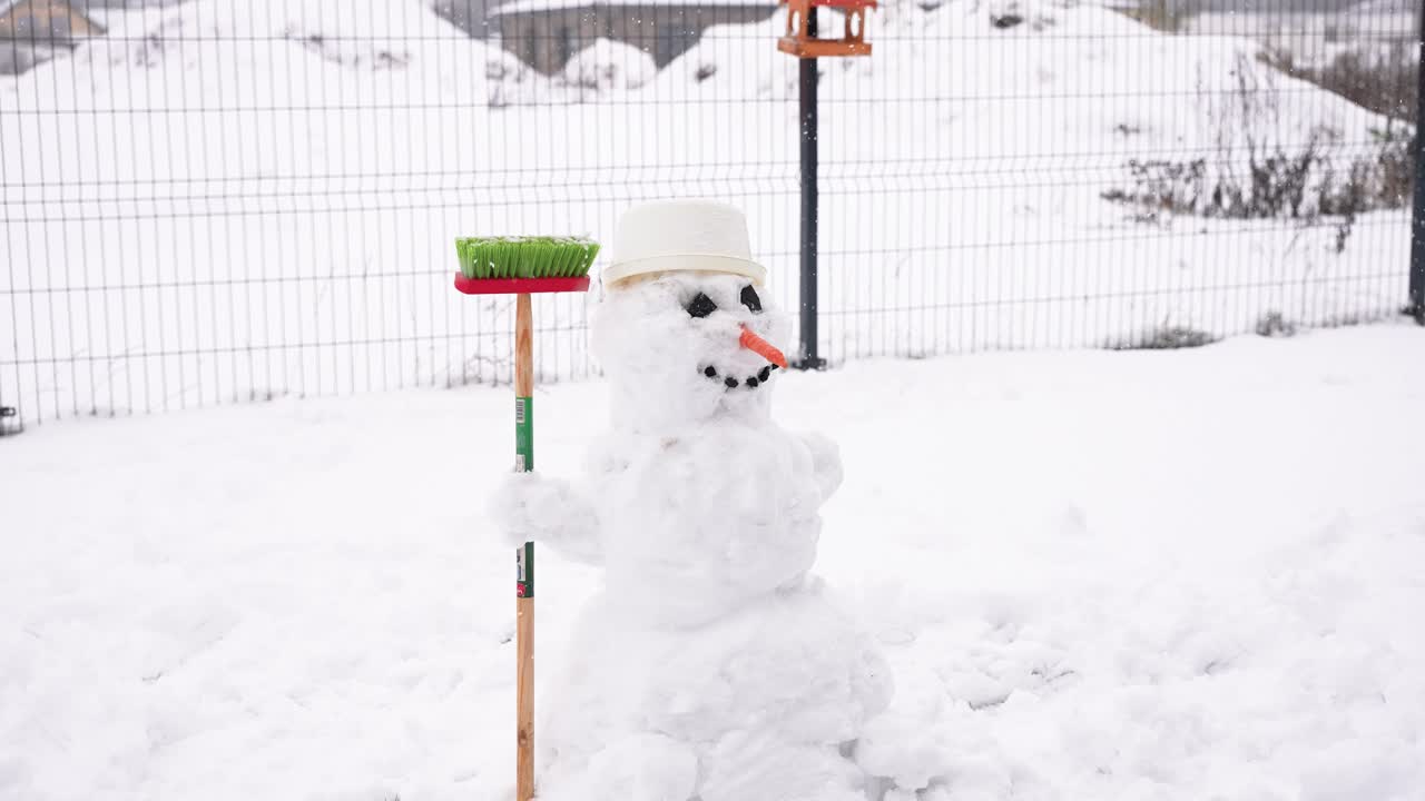 A delightful snowman stands in a snowy yard, adorned with a hat and holding a broom, conveying a playful, wintry charm. Perfectly captures winter's whimsical essence