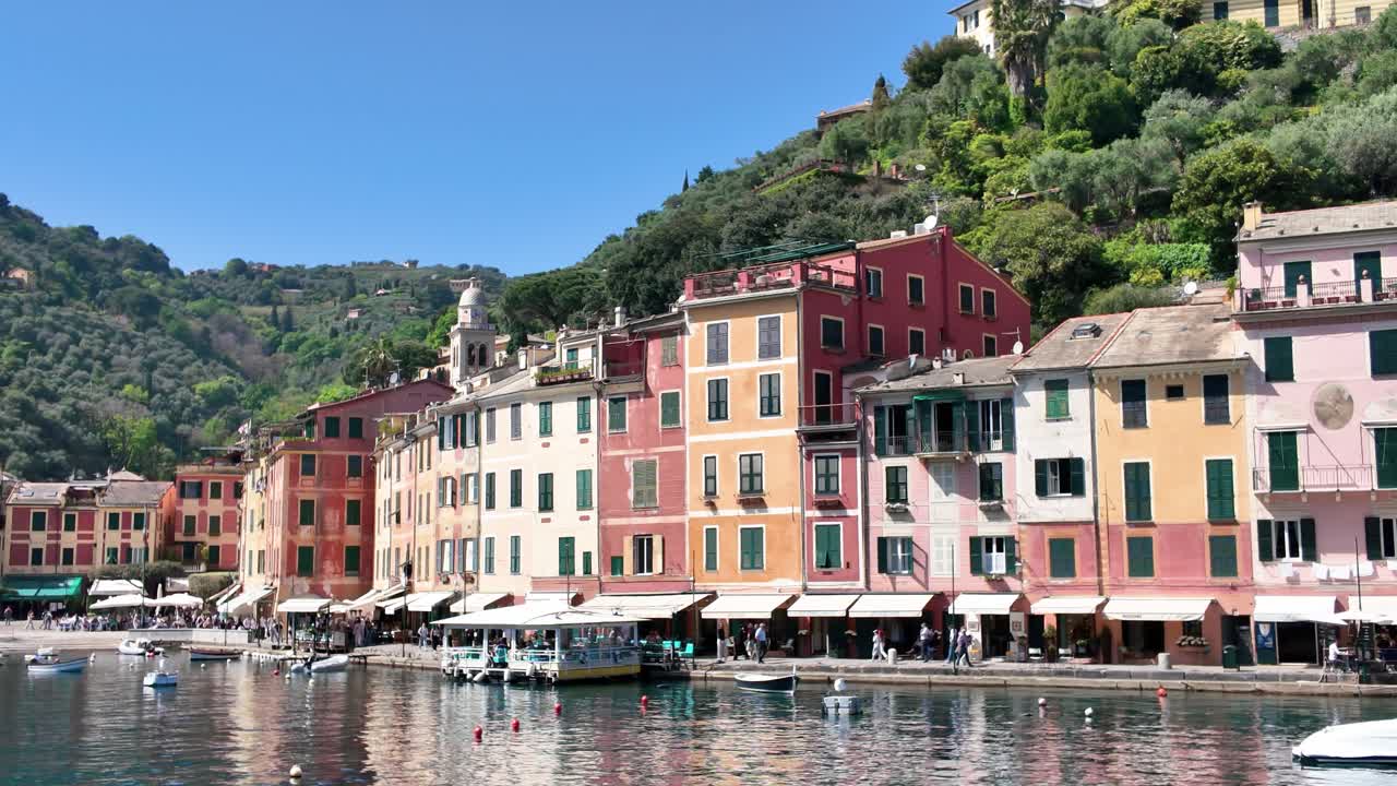 Riprese grandangolari catturano gli iconici edifici pastello sul lungomare di Portofino, il porto scintillante con barche, le rigogliose colline verdi e la luminosa luce solare mediterranea in un vivace e animato villaggio italiano.