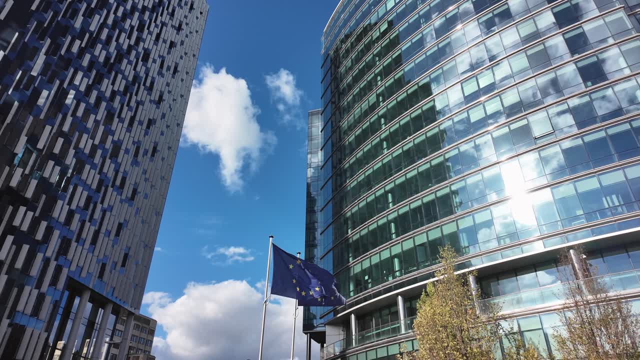 European Union flag fluttering between modern office towers near Maalbeek in Brussels, with the Lex building visible in the EU district