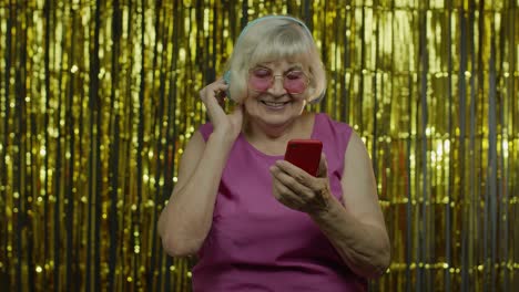 Stylish-happy-senior-old-woman-holding-cell-phone,-listening-to-music-in-headphones-and-dancing