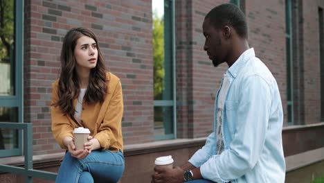 estudiantes caucásicos y afroamericanos hablando y bebiendo café sentados en la calle cerca de la universidad