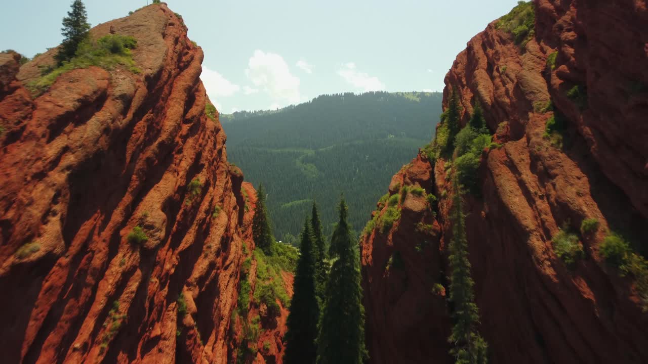 화창한 날 붉은 산을 날아가는 드론 영상, 울퉁불퉁한 봉우리, 나무가 무성한 푸른 계곡, 탁 트인 풍경, 지역의 자연미를 포착