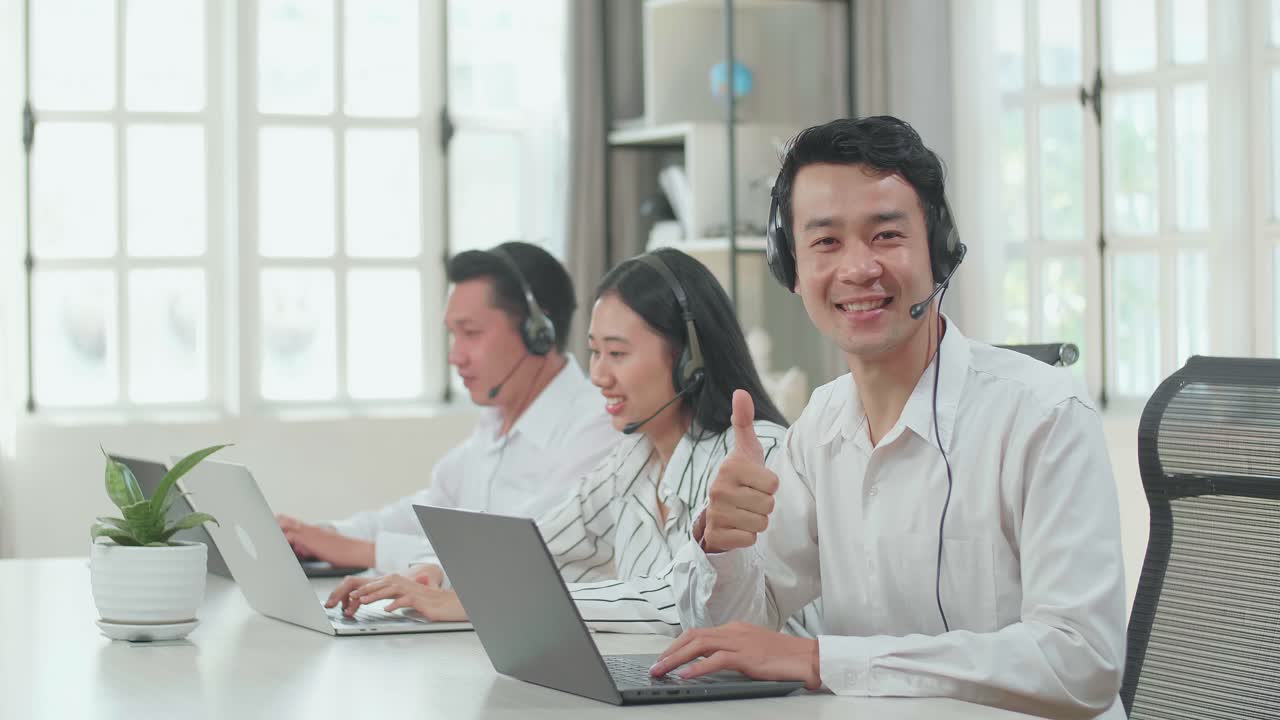 A Man Of Three Asian Call Center Agents Wearing Headset And Smile To Camera Then Thumbs Up While Two Of His Colleagues Are Speaking And Typing During The Call With Customer At The Office