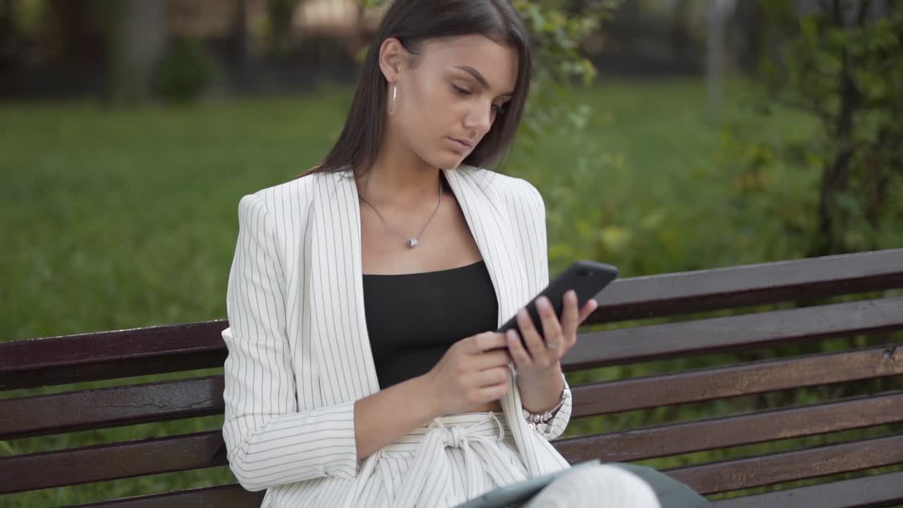 joven mujer de negocios revisa su teléfono inteligente, sentada en un banco en el parque