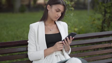 young business woman checks her smartphone, sitting on a bench in the park