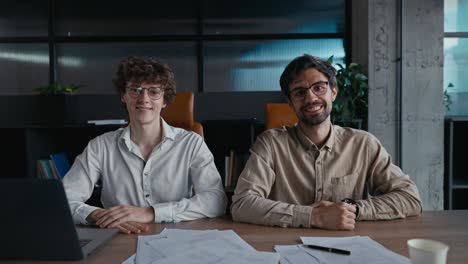 Portrait-of-a-young-guy-with-curly-hair-wearing-glasses-in-a-white-shirt-together-with-his-brunette-male-colleague-at-a-table-in-the-office