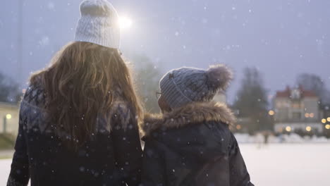 medium shot of a young mother and her daughter kin walking together to the ice skating rink for some ice skating and talking happily with each-other on a cloudy cold winter snowy evening warm clothes