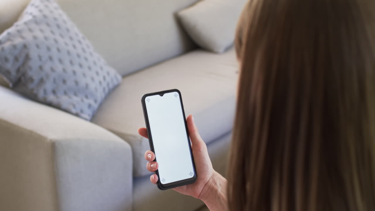 Una mujer caucásica sostiene un smartphone con la pantalla en blanco en el sofá de casa, con espacio para texto.