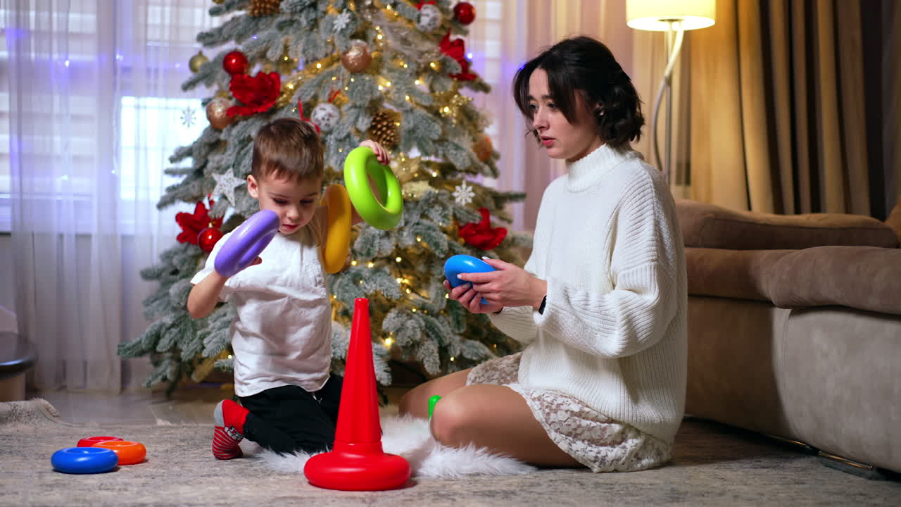 Brunette woman in white sweater plays with a kid. Mom and son build a pyramid near the Christmas tree.