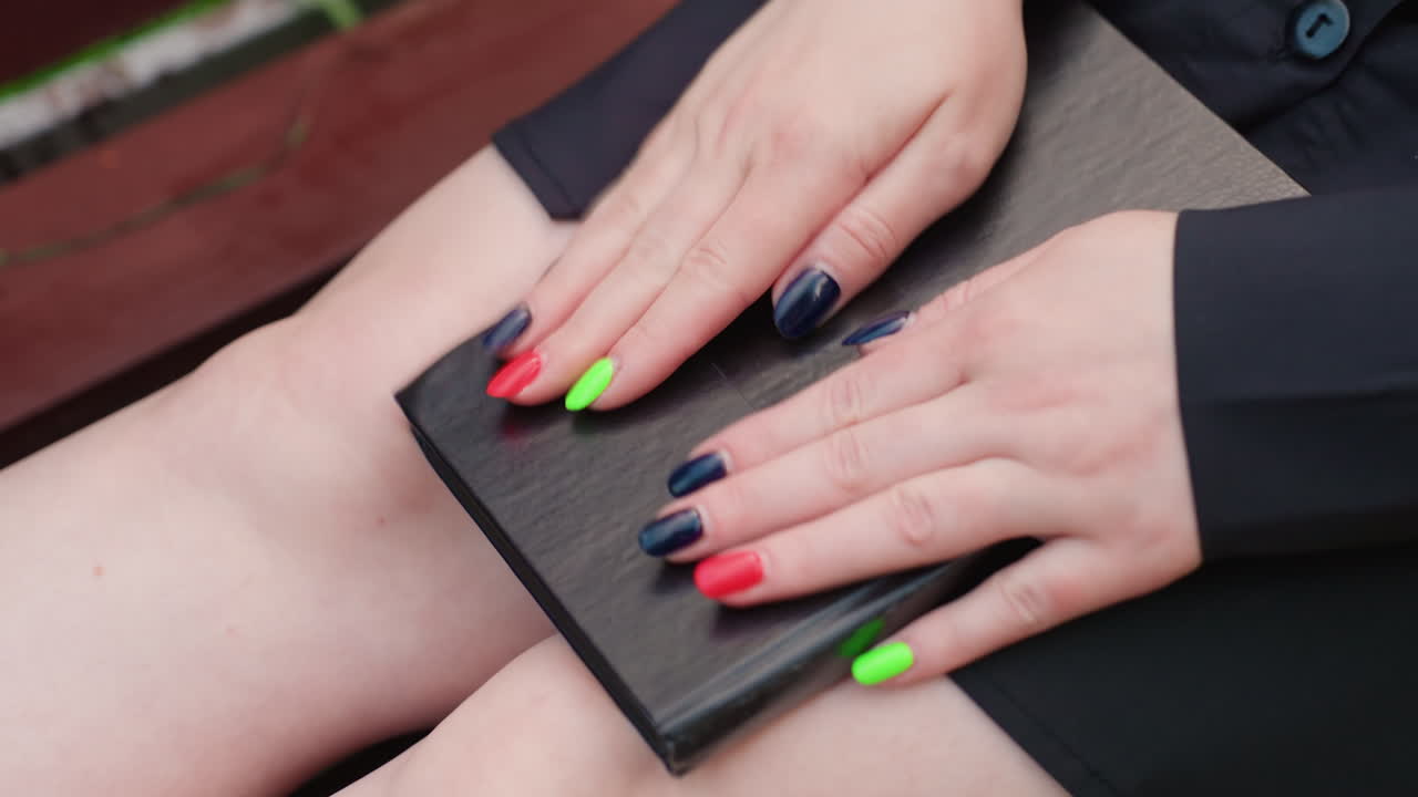 Mujer caucásica con las manos sobre un diario, manicura neón sobre una cubierta de cuero negro apoyada en el regazo, dedos trazando la textura, banco de madera, quietud sombría, recuerdo íntimo y tristeza silenciosa