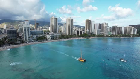 a drone pulls back showing a 4k aerial view of a stunning tropical vacation destination full of families on the beach, surfers surfing and couples enjoying a romantic dinner cruise