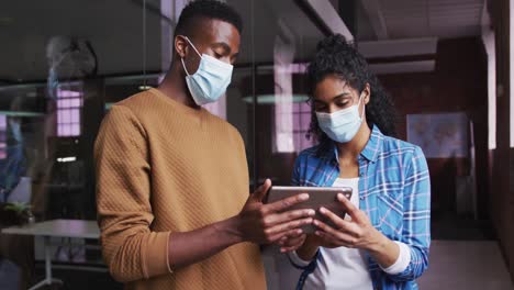 Diverse-female-and-male-business-colleagues-wearing-masks-in-discussion-looking-at-digital-tablet