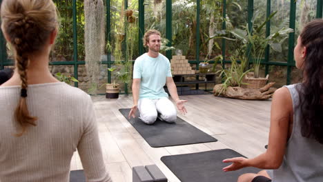 leading yoga class, instructor kneeling on mat in glass house with students