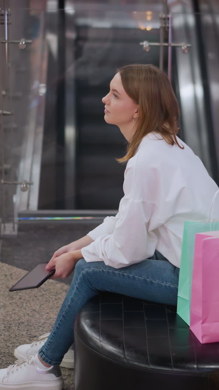 Young woman seated on bench in shopping mall holding tablet surrounded by festive decorations, illuminated star decor, and shopping bags, with escalators in background