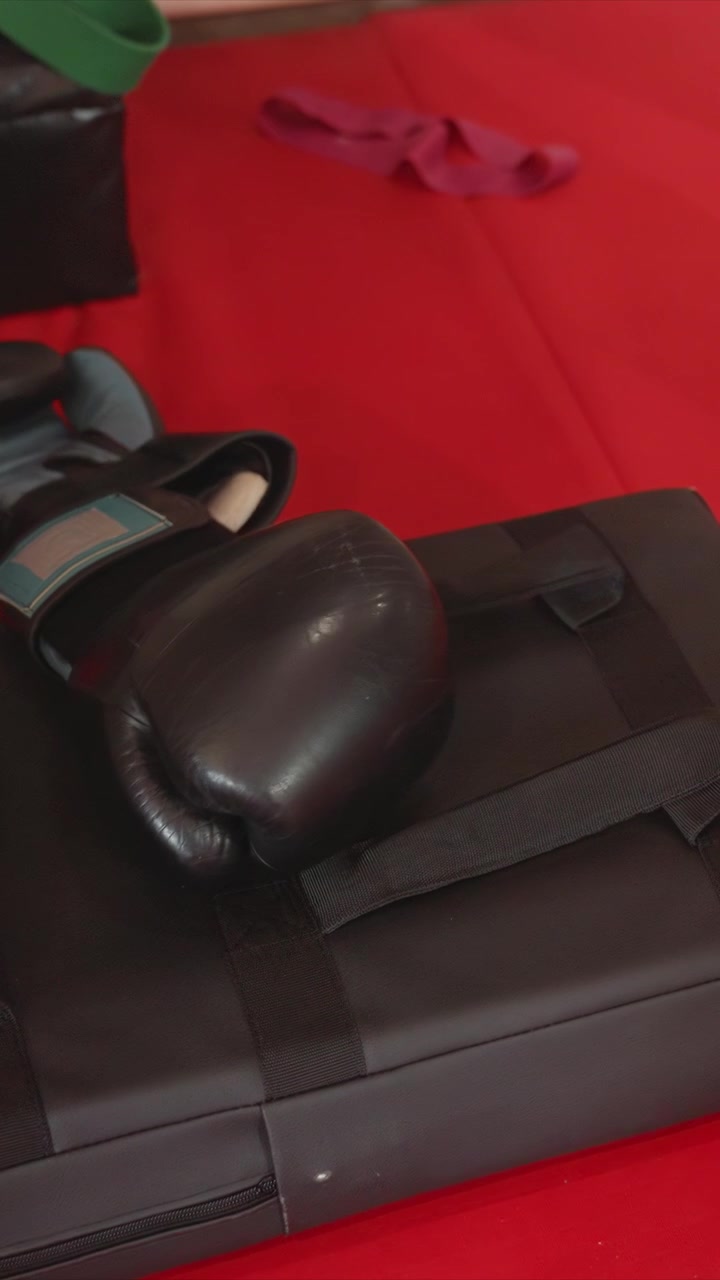 Primer plano de guantes de boxeo negros descansando sobre un saco acolchado. Los guantes muestran marcas de uso y detalles de textura. Una colchoneta roja proporciona un fondo consistente en toda la escena.