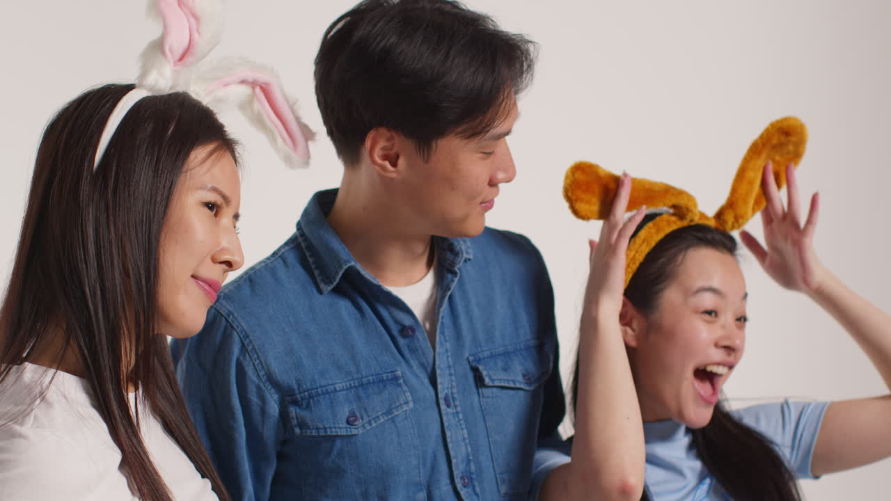 색 스튜디오 배경 앞의 젊은 친구 그룹이 가짜 동물 귀로 사진 부스 스타일의 초상화에 포즈를 취하는 재미를 누리고 있습니다.