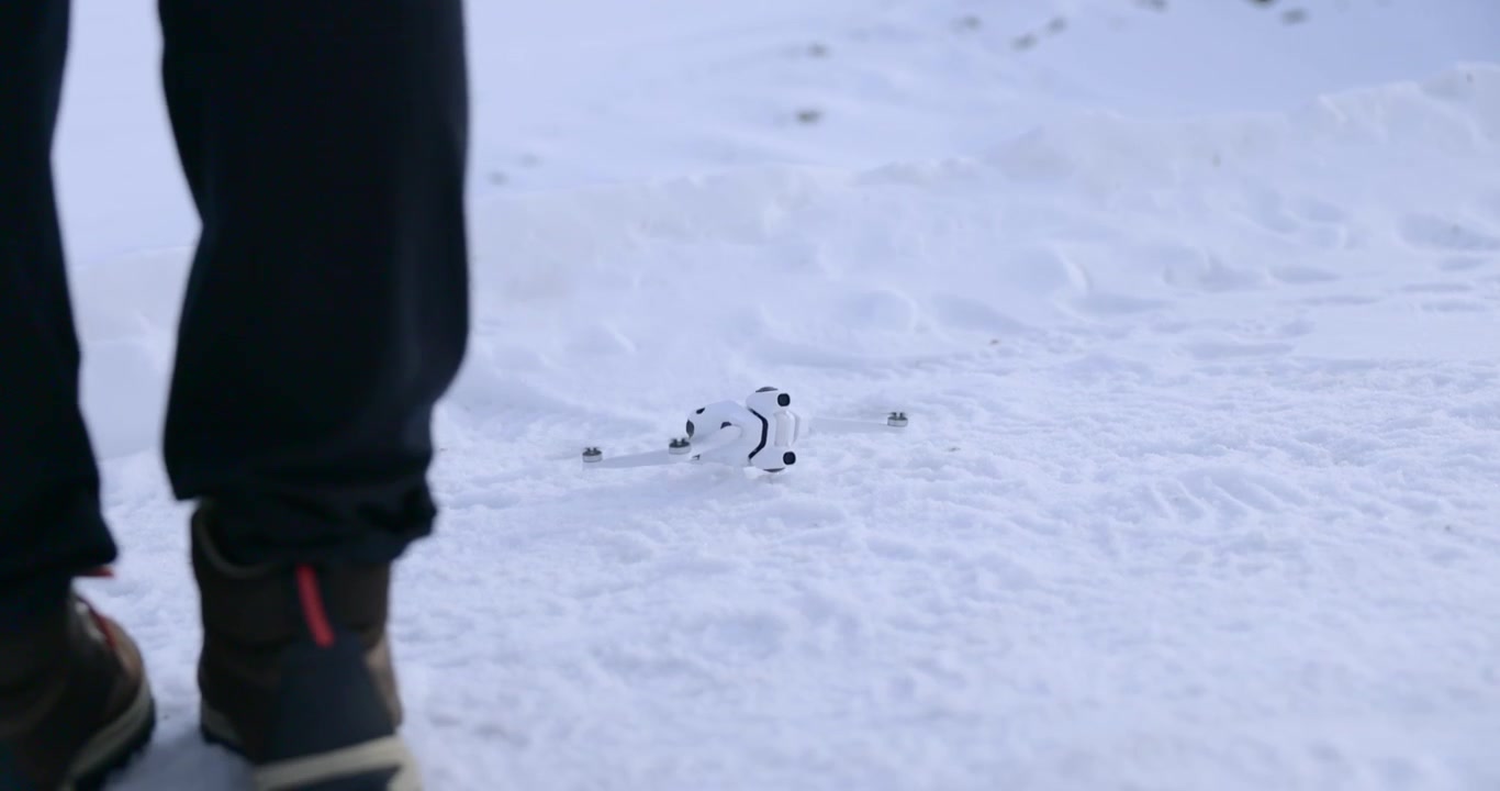 Vista ravvicinata di un piccolo drone quadricottero bianco che atterra sulla neve fredda accanto alle gambe del pilota all'aperto.