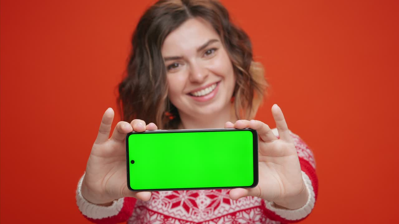 navidad joven mujer sorprendida sosteniendo teléfono inteligente con pantalla verde dedo índice mostrando gesto de deslizamiento en ropa de navidad para copiar espacio para anunciar sus productos en fondo rojo. concepto de venta