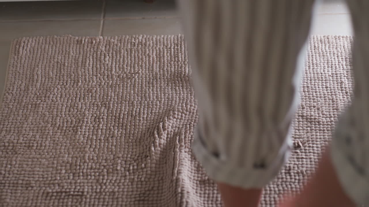 High angle view shows young woman legs setting on soft bath mat, gently drying feet after bath, water glistening on skin, hygiene moment on tiled floor with absorbent textured rug ensuring comfort