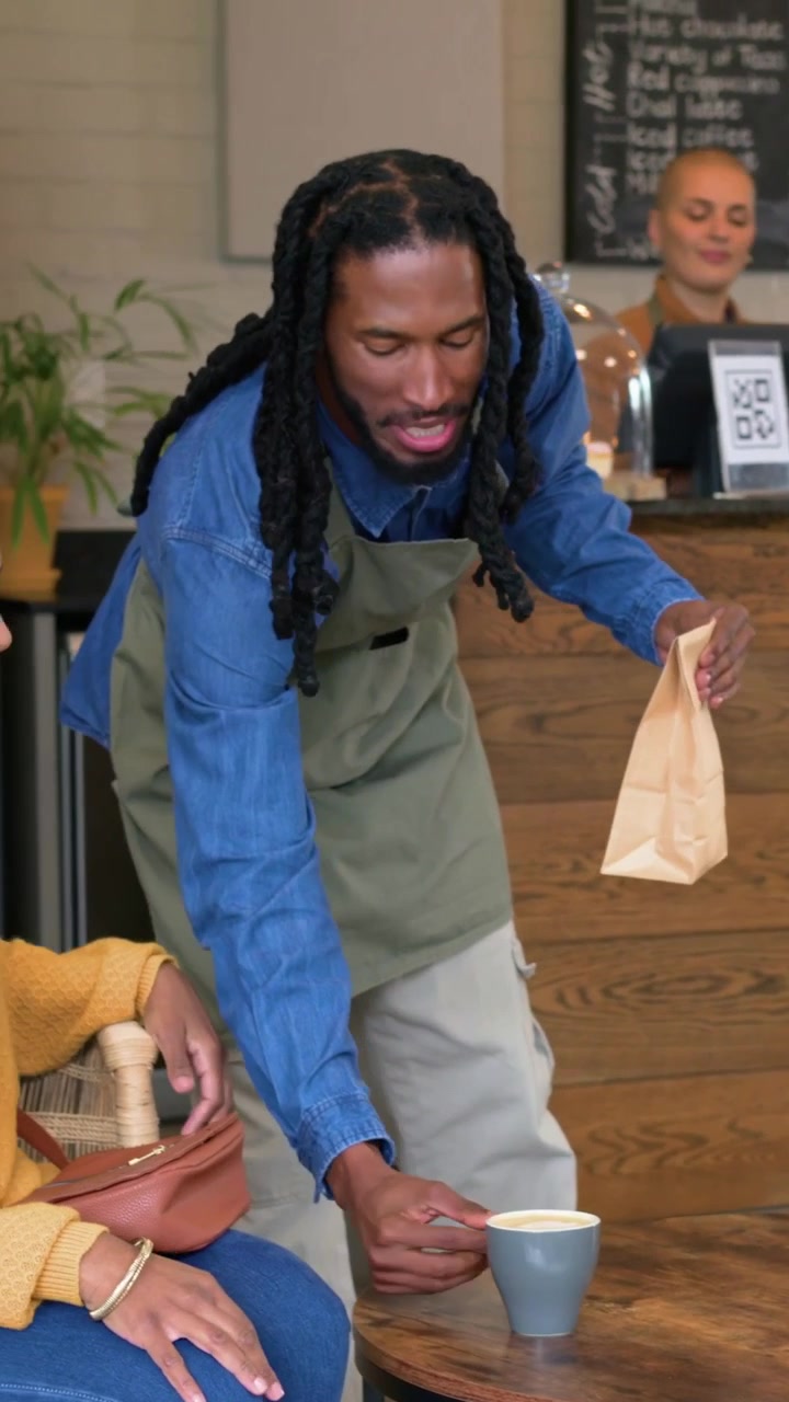 Vertical video: Placing mug and folding paper bag barista serving woman wearing yellow top in cafe
