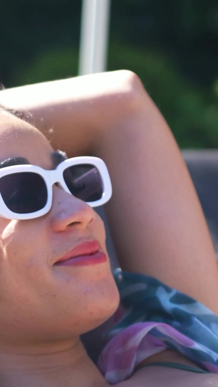 Vertical video: Panning woman on chair in green swimsuit stretching scarf shifting relaxing pool