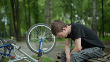 un niño blanco con una camiseta negra se sienta pensativo en un banco del parque con la cabeza hacia abajo, su bicicleta está al revés a su lado, sus neumáticos todavía girando, y el fondo presenta una vista borrosa de árboles