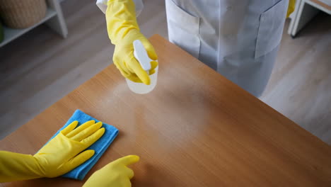 person in yellow gloves cleaning a wooden table with a spray bottle and cloth