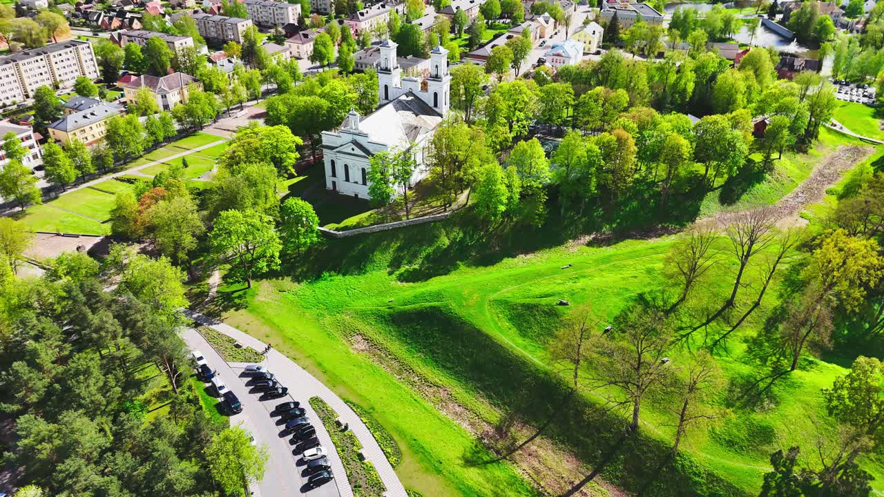 A white church sits above a sunlit park with winding roads, rolling green lawns, and surrounding forest near a quiet residential district in a European city.