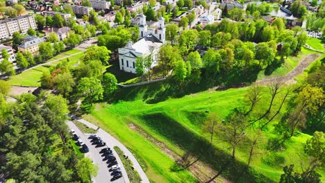 a white church sits above a sunlit park with winding roads, rolling green lawns, and surrounding forest near a quiet residential district in a european city.