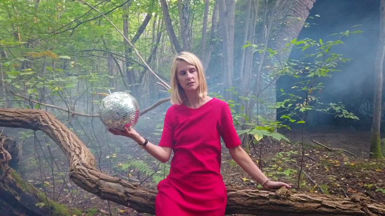 Perfect slow motion cinematic shot of a sexy blond girl with a disco ball in a read dress lonely in russian forest sitting on a tree Foto shoot on lost places in nature 1080, 24p by Philipp Marnitz