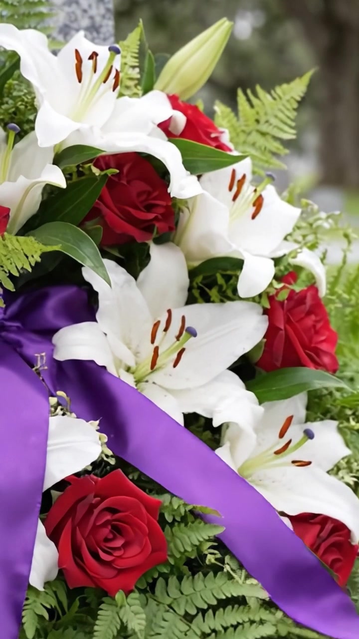 Una splendida composizione floreale di rose rosse e gigli bianchi con nastri viola, che simboleggia il ricordo in un tranquillo ambiente cimiteriale.