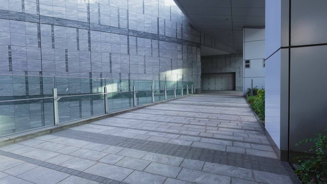 A modern urban walkway with a glass railing, leading to an entryway with a large, textured wall