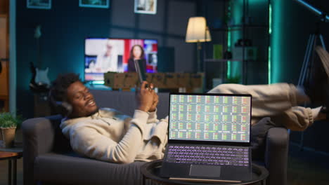 man enjoying music and reading e books on tablet while waiting for stock market financial figures to get updated on laptop. person at home next to pc showing trading investment graphs. camera b.