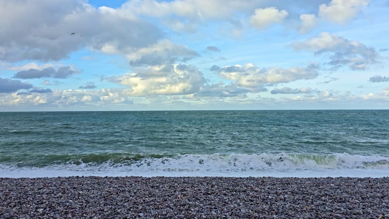 Wide static shot of calm sea and cloudy sky over a pebble beach in Normandy France with natural coastal atmosphere