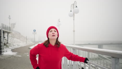 entrenadora haciendo ejercicio al aire libre durante el invierno, sosteniendo una barandilla de hierro para el equilibrio mientras estira la pierna hacia adelante y hacia atrás, rodeada de tierra nevada, atmósfera brumosa, río lejano, puente en el fondo